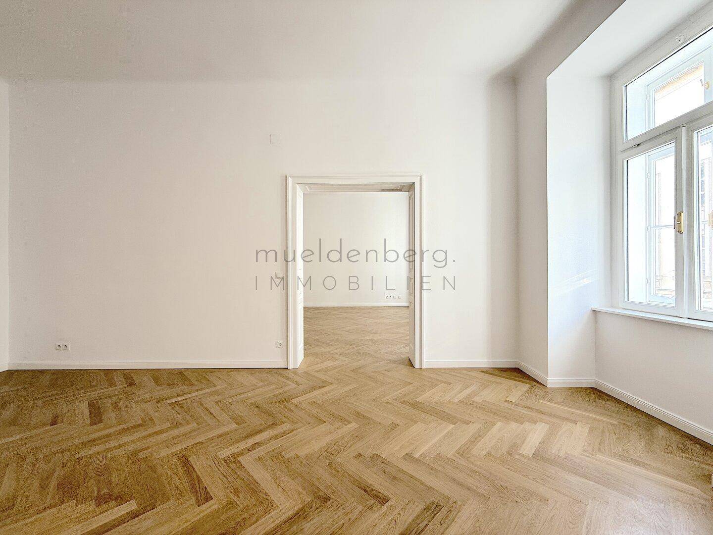 Heller Raum mit Fischgrätparkett und einem großen Fenster, das viel Tageslicht hereinlässt.