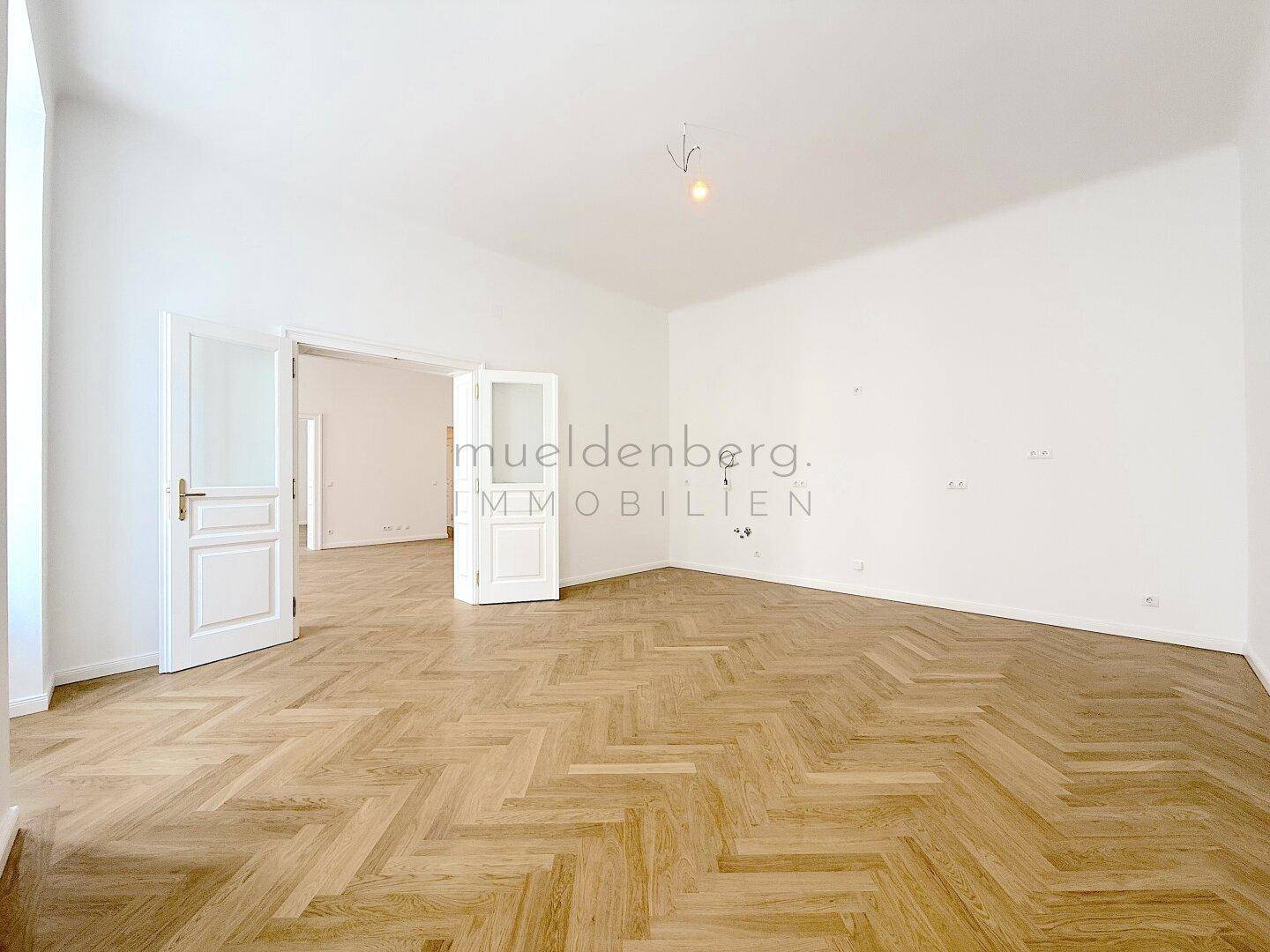 Geräumiger, heller Raum mit Fischgrätparkett und Doppeltüren, die zu einem weiteren Zimmer führen.