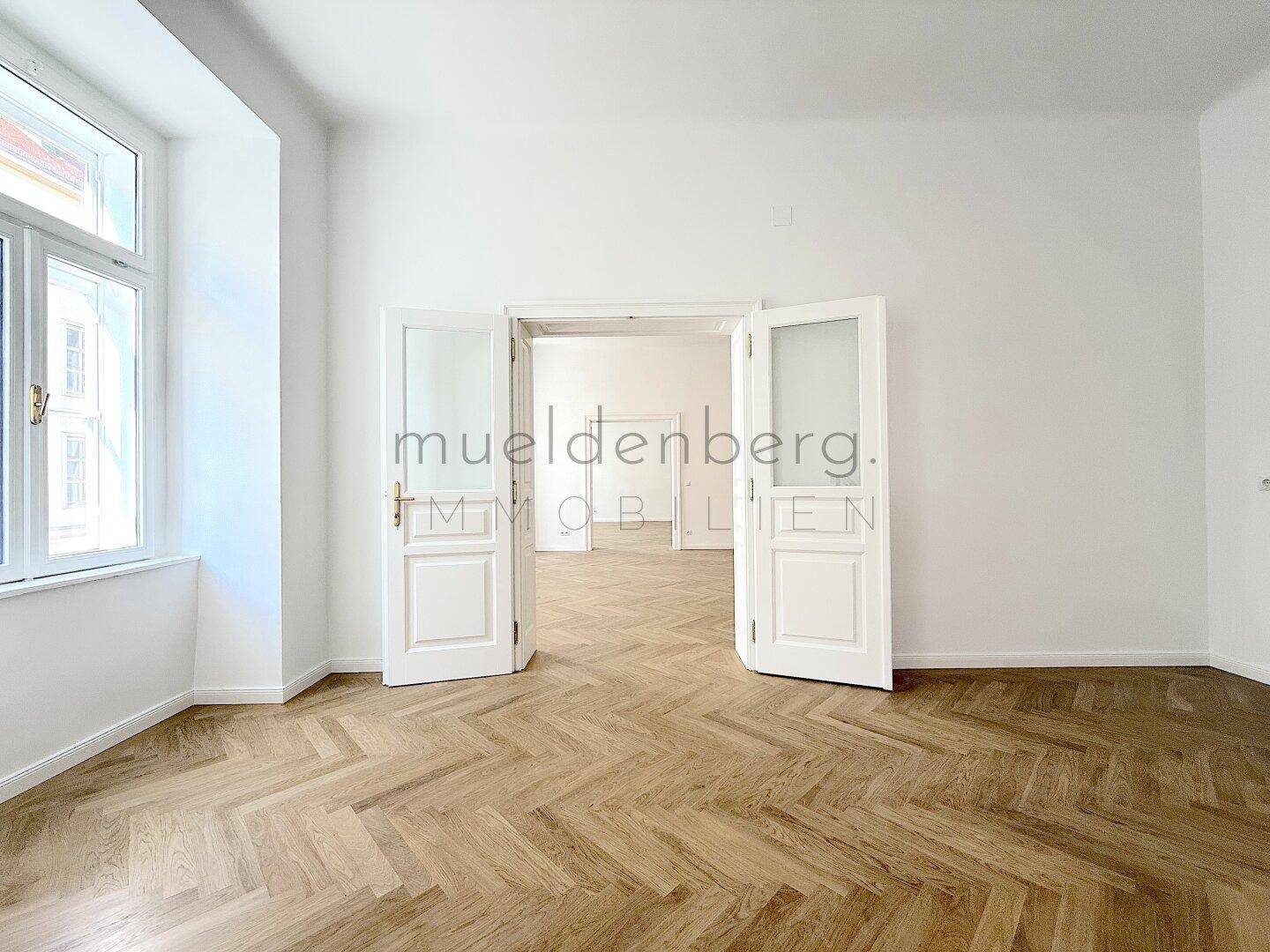 Heller Raum mit Fischgrätparkett und Blick in angrenzende Zimmer.