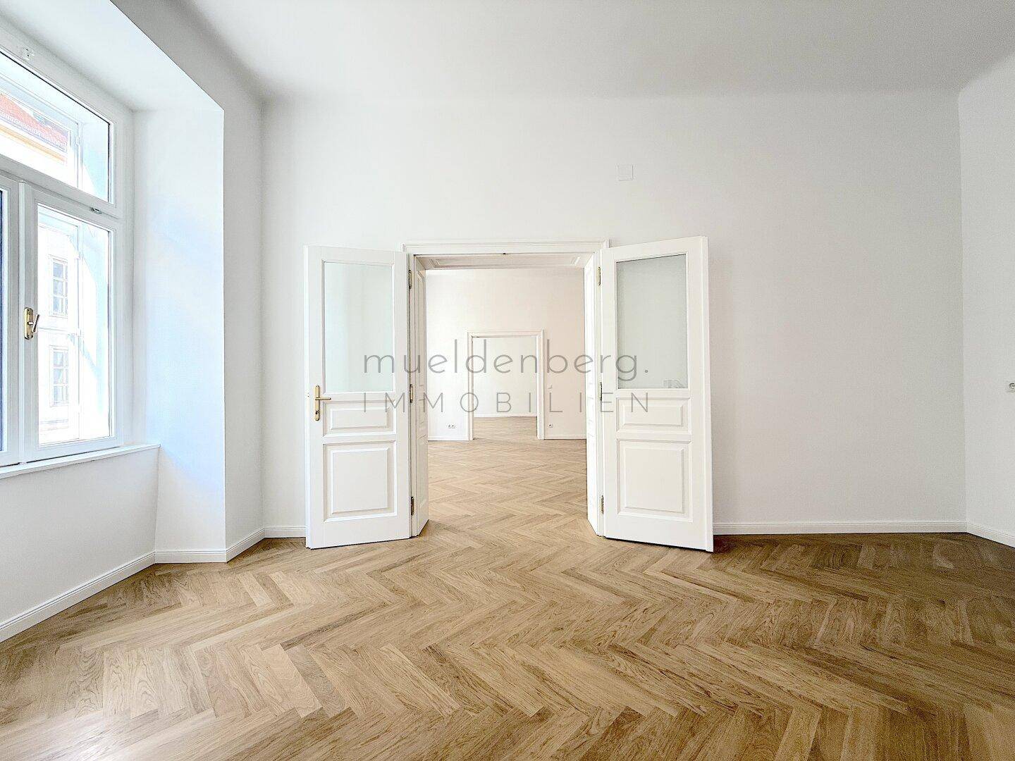 Heller Raum mit Fischgrätparkett, großem Fenster und Doppeltüren, die zu einem weiteren Zimmer führen.
