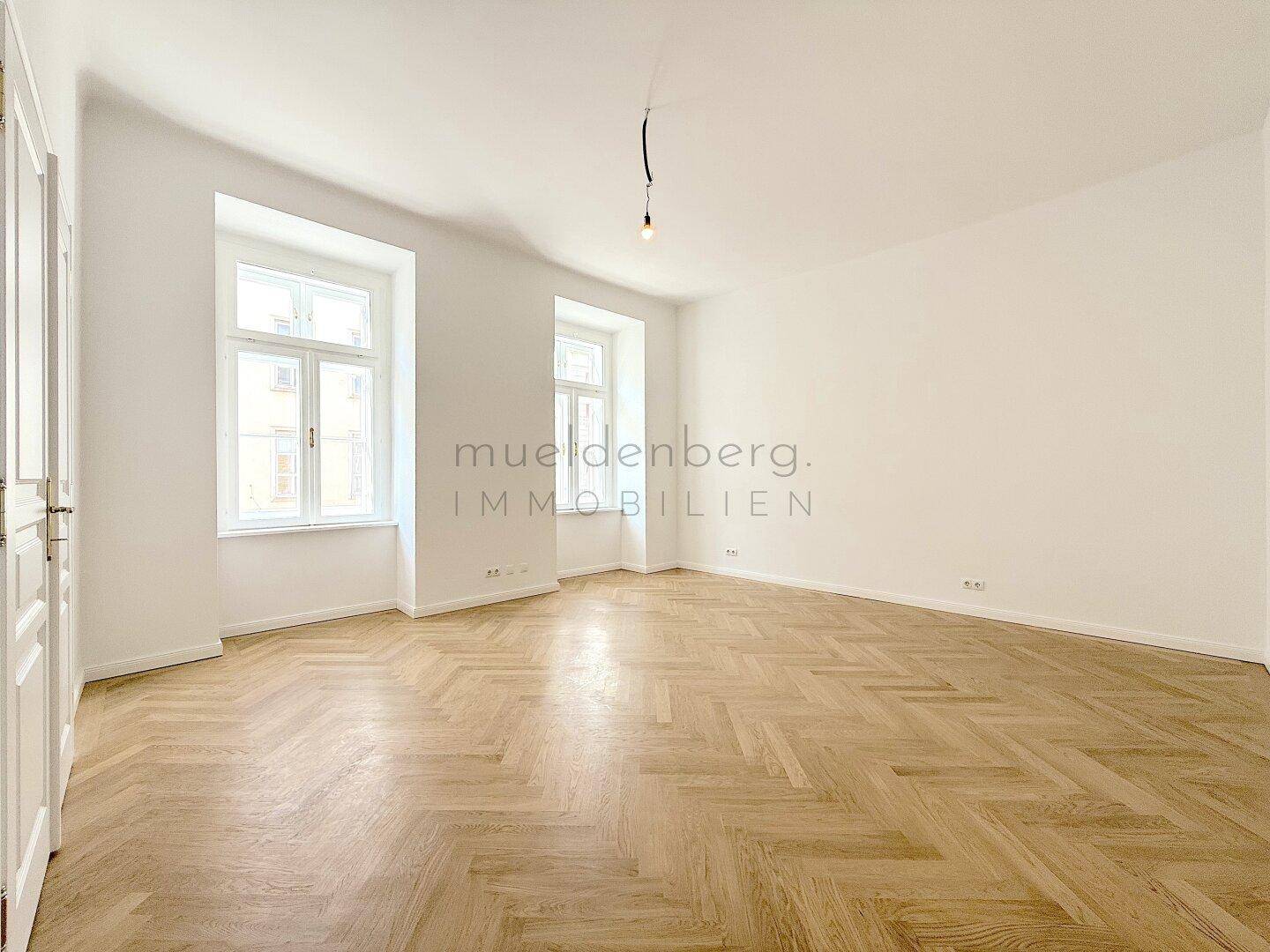 Geräumiges Zimmer mit zwei hohen Fenstern und elegantem Fischgrätparkett, ideal für Wohnzwecke.