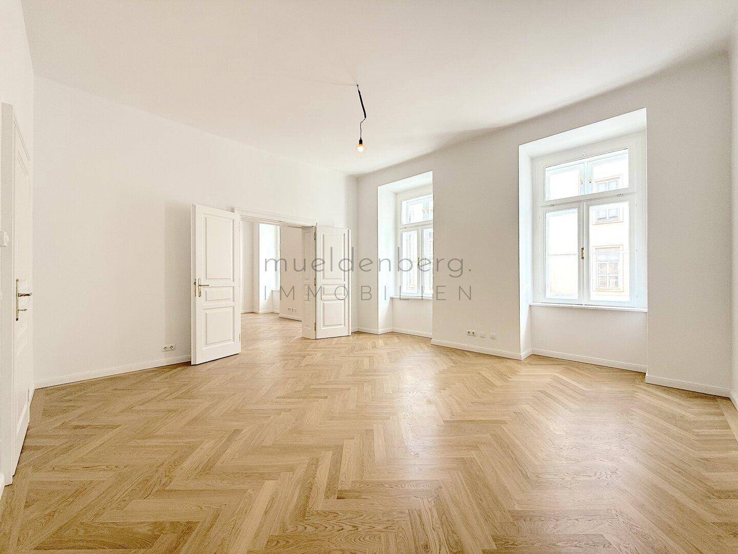 Großzügiges Zimmer mit Fischgrätparkett und Zugang zu weiteren Räumen, lichtdurchflutet und offen.