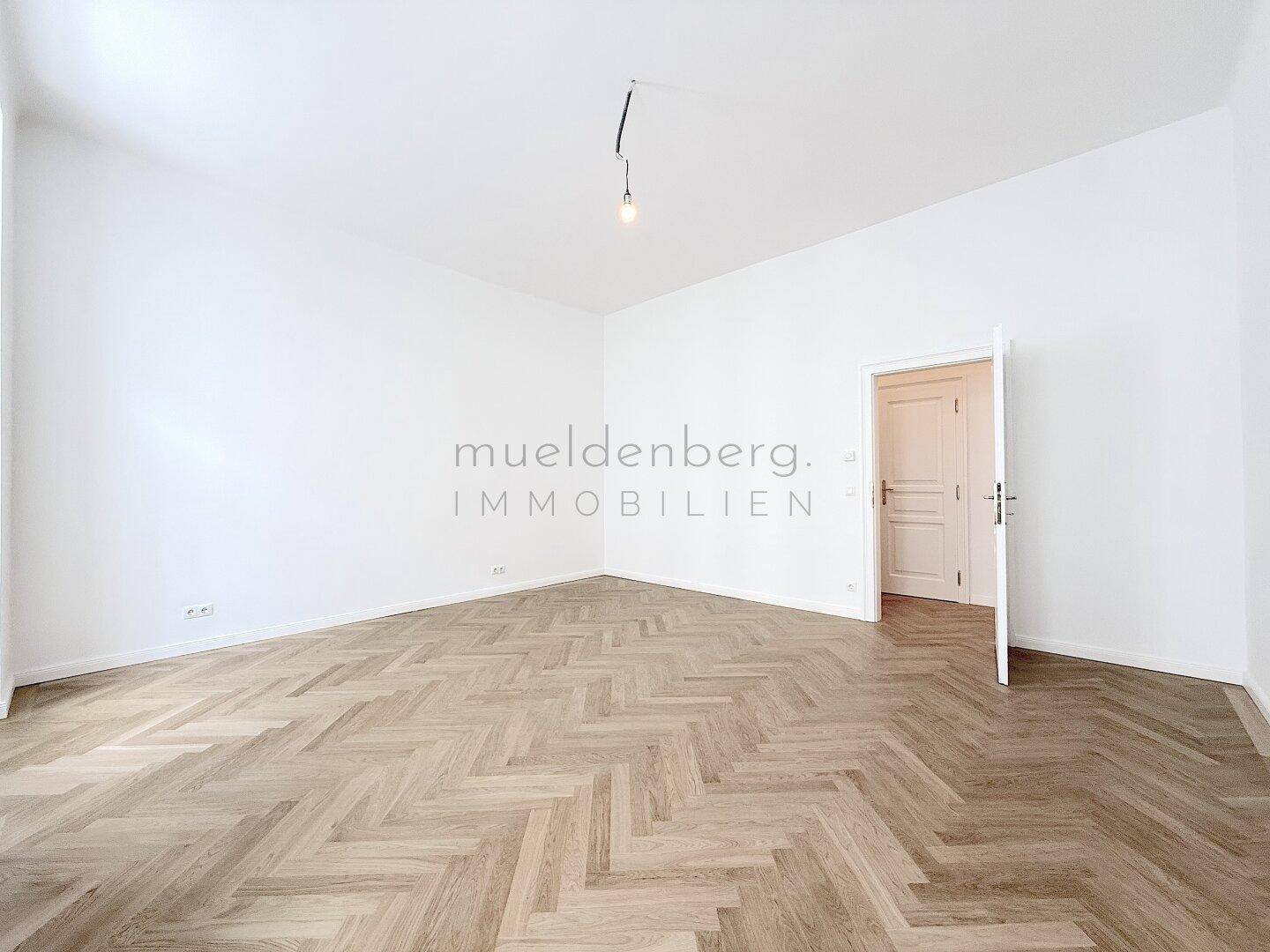 Leeres Zimmer mit Fischgrätparkett und einer offenen Tür, die zu einem weiteren Bereich führt.