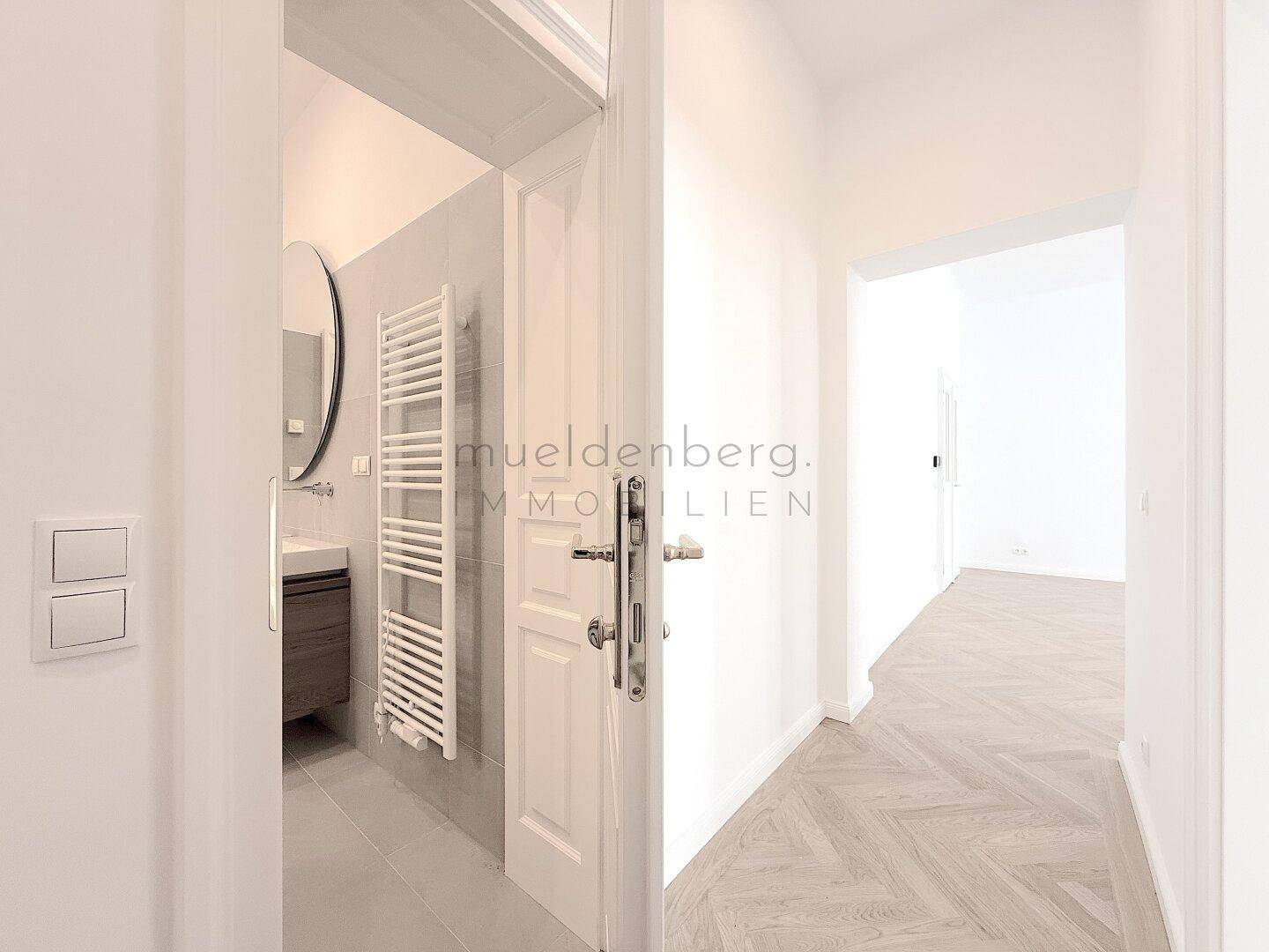Flur mit Fischgrätparkett und Blick in ein modernes Badezimmer mit Handtuchheizkörper und Spiegel.