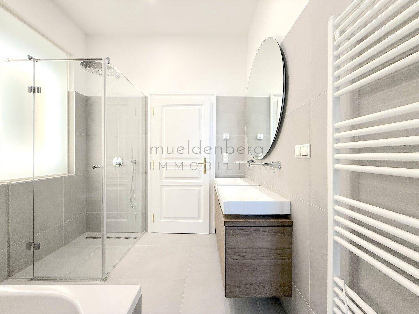 Modernes Badezimmer mit großer Dusche, Waschtisch und ovalem Spiegel, in hellen Grautönen gehalten.