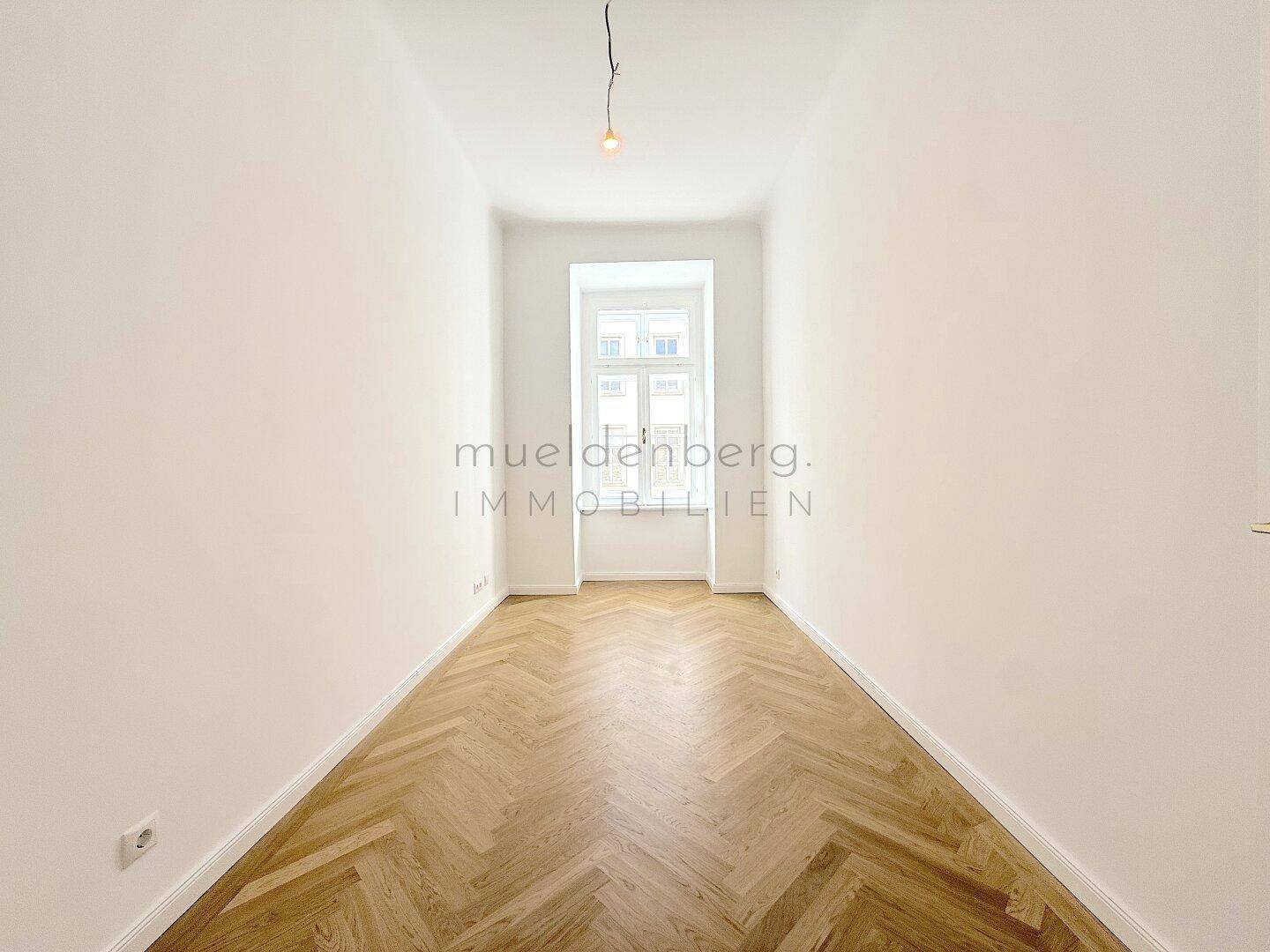 Schmaler, heller Raum mit Fischgrätparkett und einem hohen Fenster, ideal als Schlafzimmer.