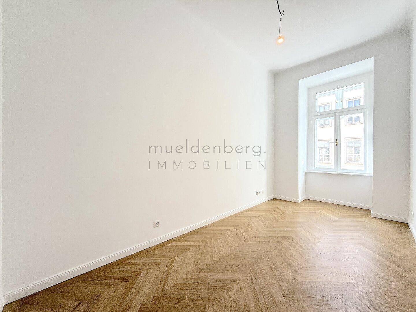 Helles Zimmer mit Fischgrätparkett und einem hohen Fenster, bietet viel Tageslicht.