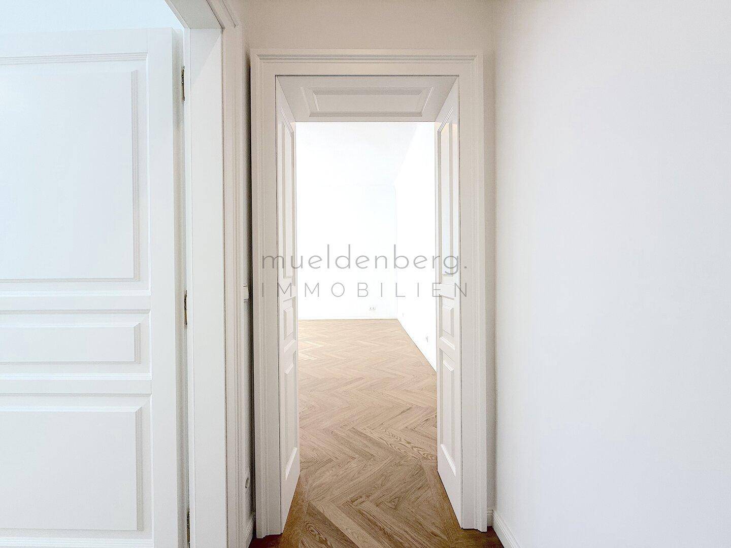 Blick durch eine Doppeltür in einen hellen Raum mit Fischgrätparkett und hohen Decken.