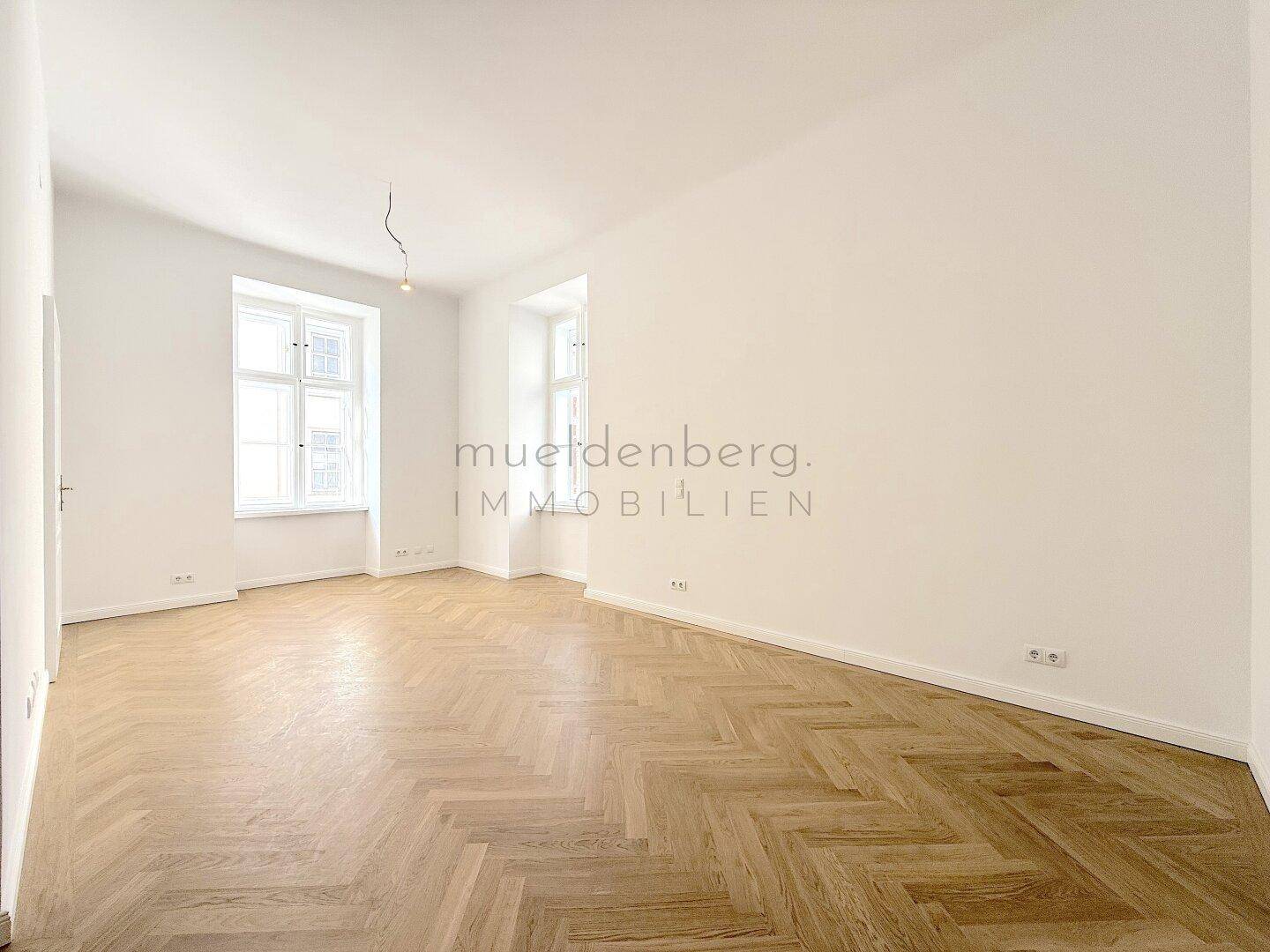 Geräumiger, heller Raum mit Fischgrätparkett und großen Fenstern, ideal für vielfältige Nutzung.