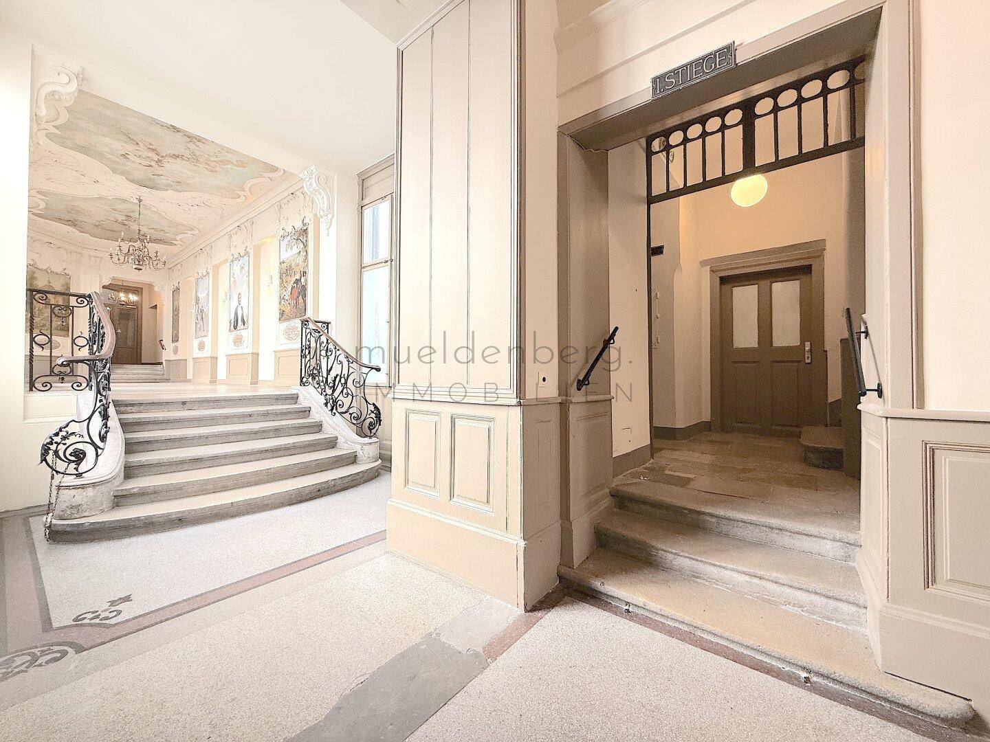 Prunkvolles Treppenhaus mit kunstvollen Geländern, Deckenmalerei und großen Fenstern im Altbau.