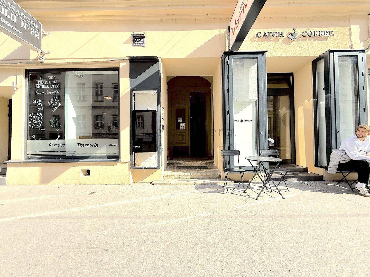 Erdgeschossfassade eines Gebäudes mit Geschäften und Cafés, belebtes Stadtbild.