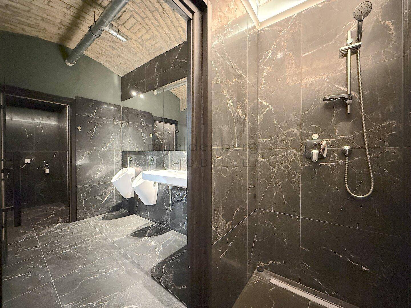 Modernes Badezimmer mit dunklen Marmorfliesen, Dusche und Blick auf Urinale und Waschbecken.