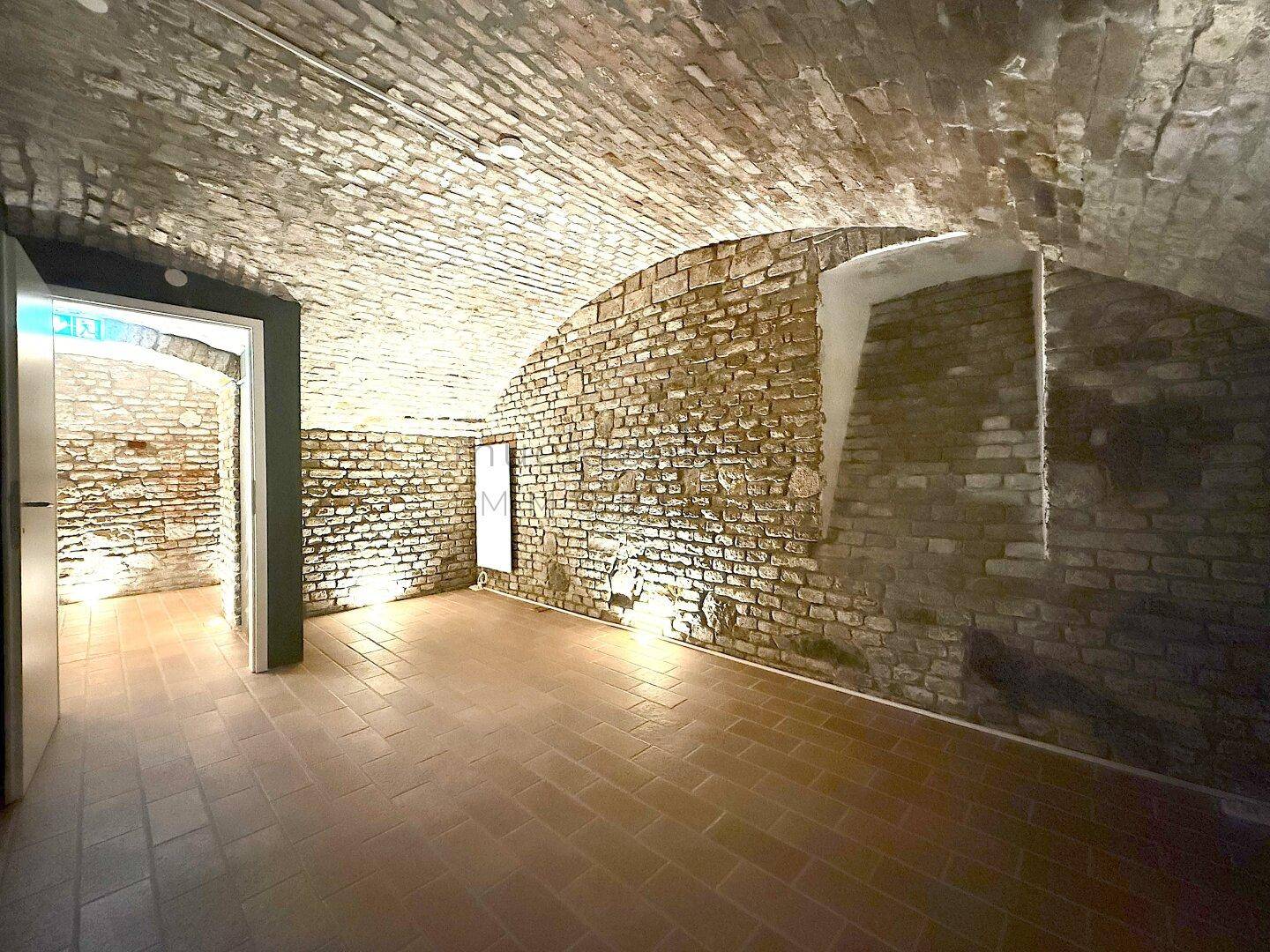 Historischer Kellerraum mit freigelegten Ziegelwänden und Gewölbedecken, beleuchtet und mit Terrakottafliesen ausgelegt.