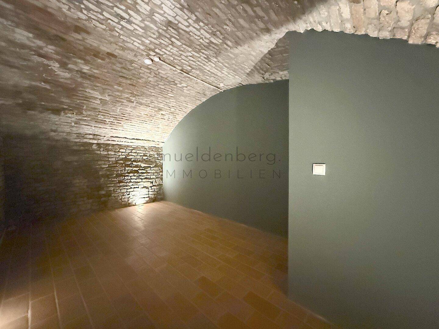 Kellerraum mit Gewölbedecke, einer geschwungenen dunkelgrünen Wand und traditionellem Terrakotta-Boden, stimmungsvoll beleuchtet.