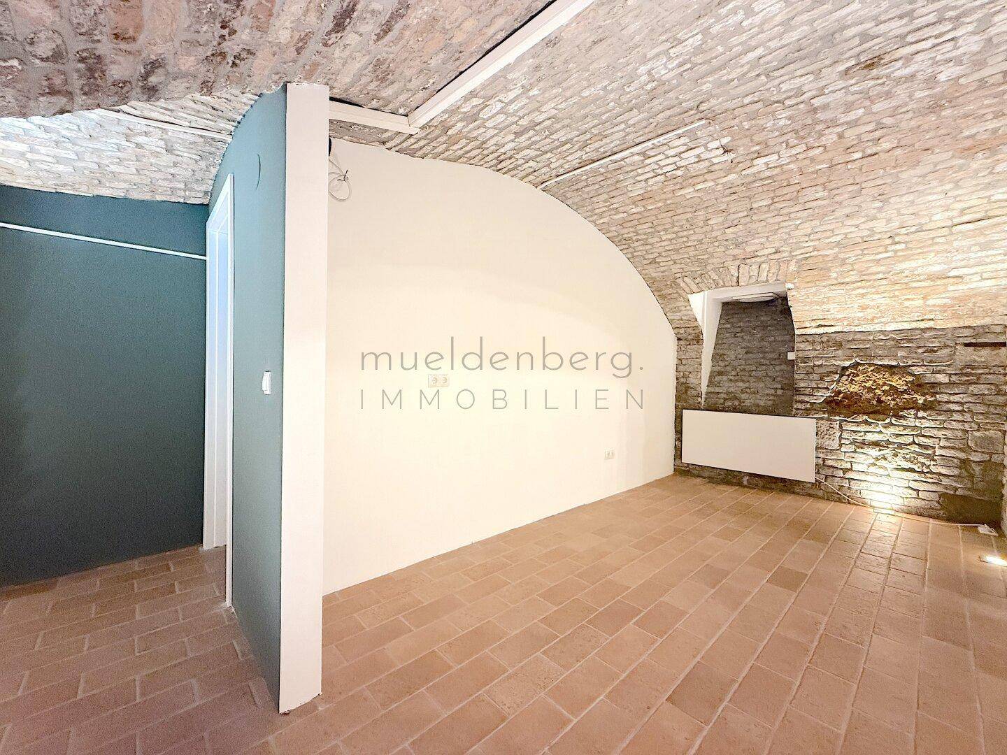 Historischer Raum mit Gewölbedecke und Ziegelwänden, ergänzt durch eine moderne Trennwand.