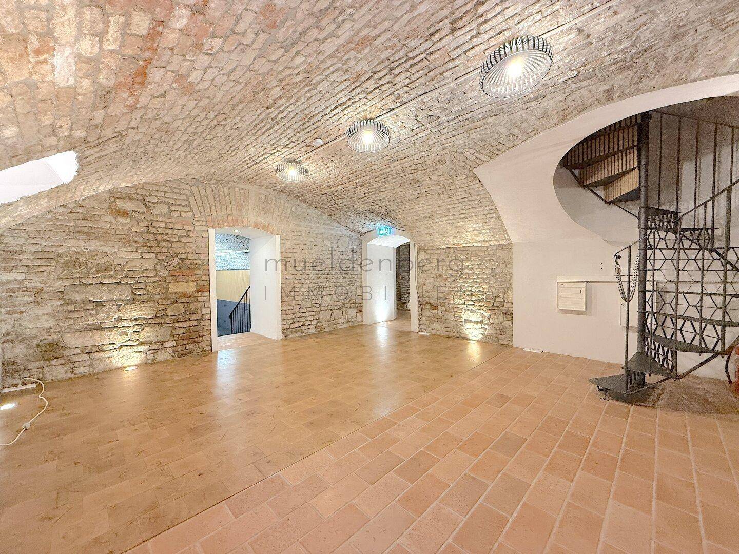 Weitläufiger Raum mit historischer Gewölbedecke, Ziegelwänden und einer markanten Wendeltreppe.