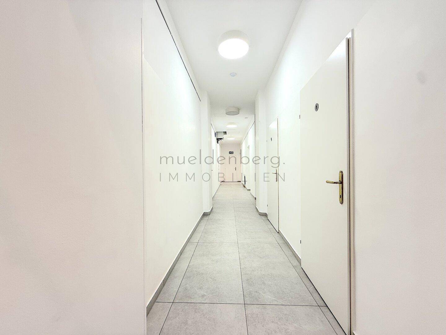 Heller, langer Flur mit weißen Wänden und mehreren Türen, ausgestattet mit modernen Deckenleuchten.