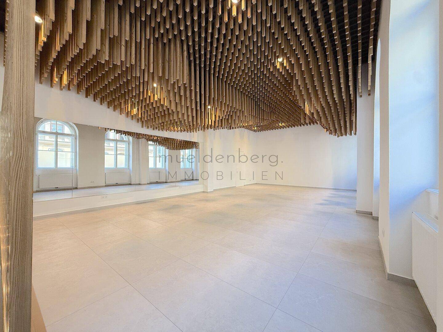 Großer, heller Gewerberaum mit Spiegelwand, Holzlamellendecke und Fliesenboden.