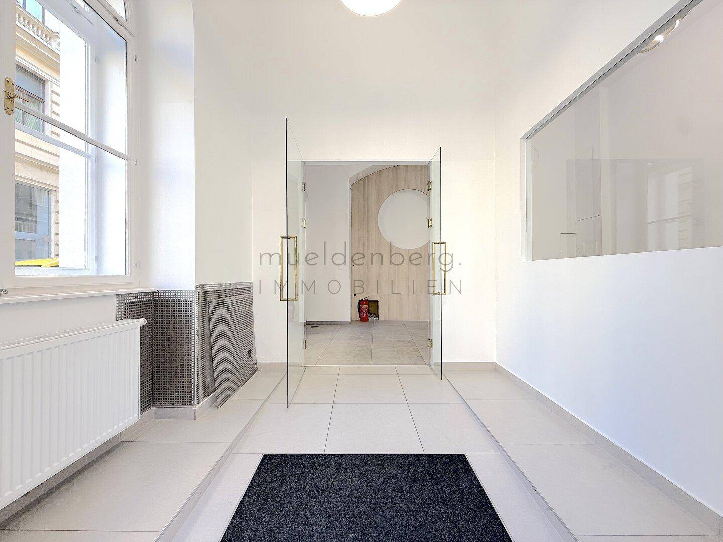 Heller Eingangsbereich mit Fliesenboden, modernen Glastüren und Blick auf den dahinterliegenden Raum.