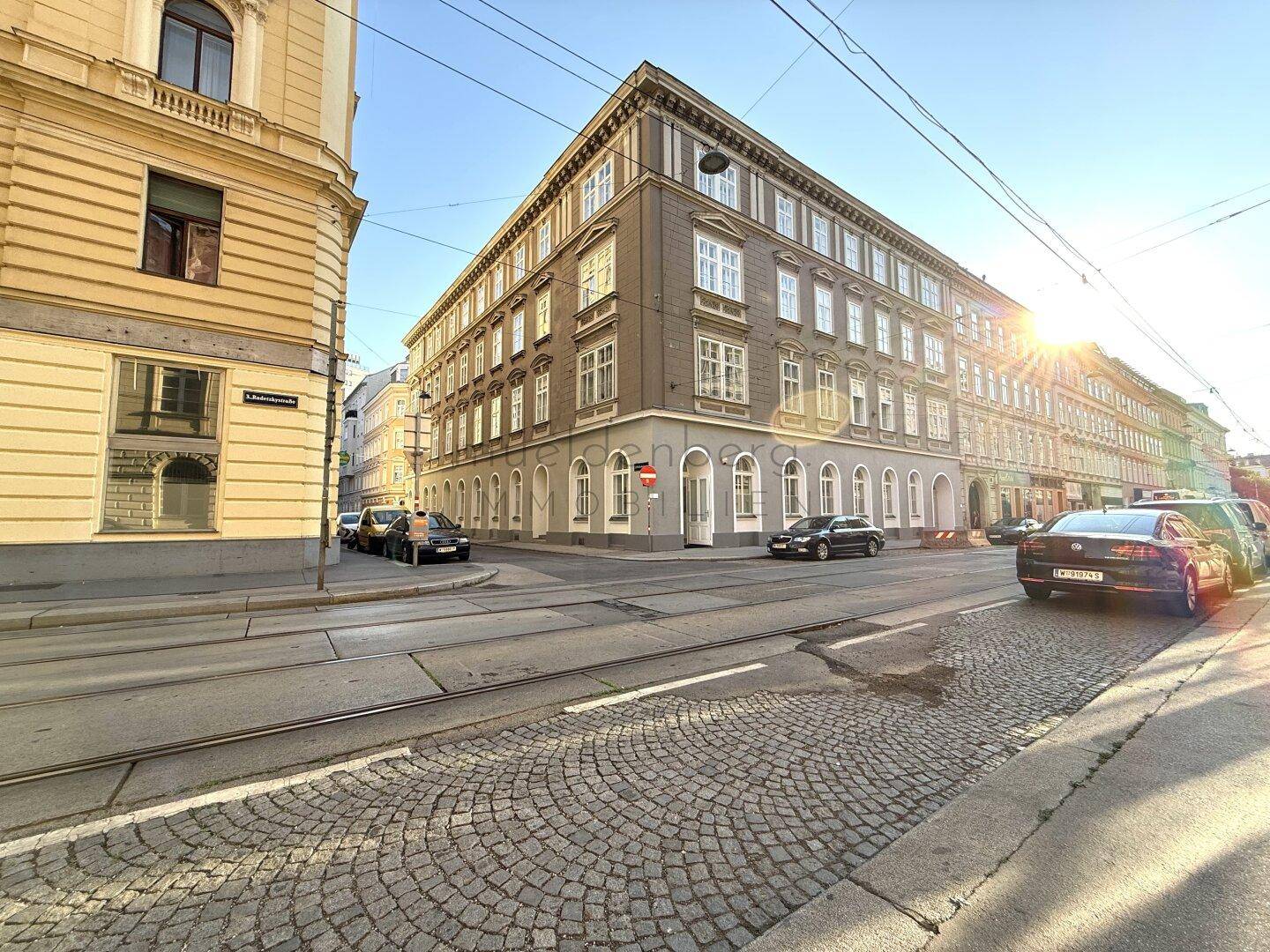 Historisches Eckgebäude an einer belebten Straße mit Kopfsteinpflaster und Straßenbahnschienen bei Sonnenuntergang.