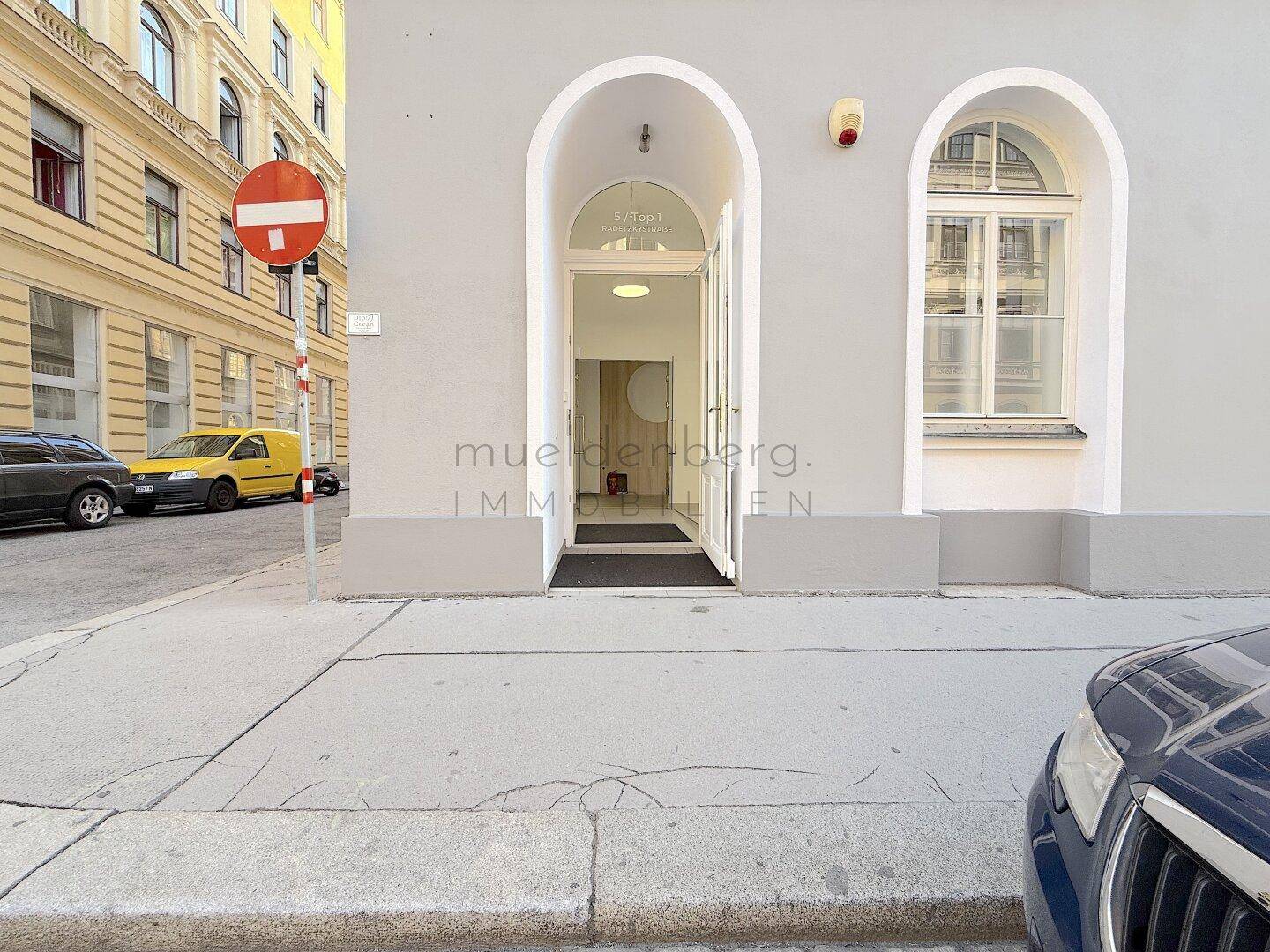 Eingangsbereich eines Gebäudes mit großen Rundbogenfenstern und einer modernen Glastür zur Straße hin.