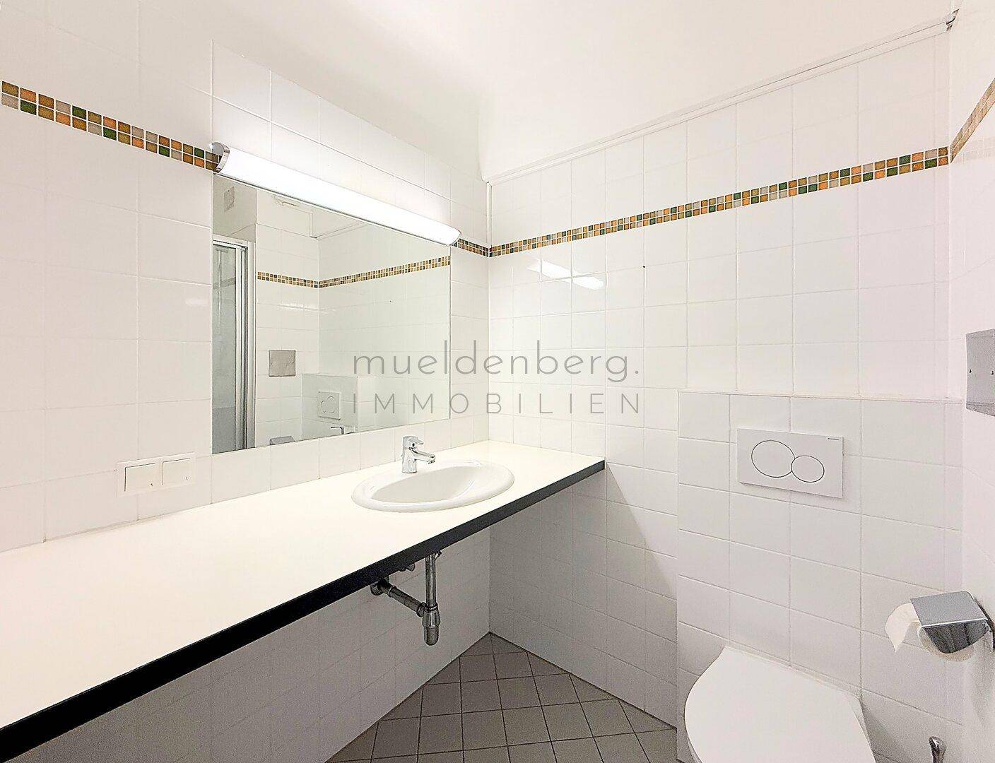 Badezimmer mit weißen Fliesen, Waschbecken, Toilette und einem großen Spiegel.