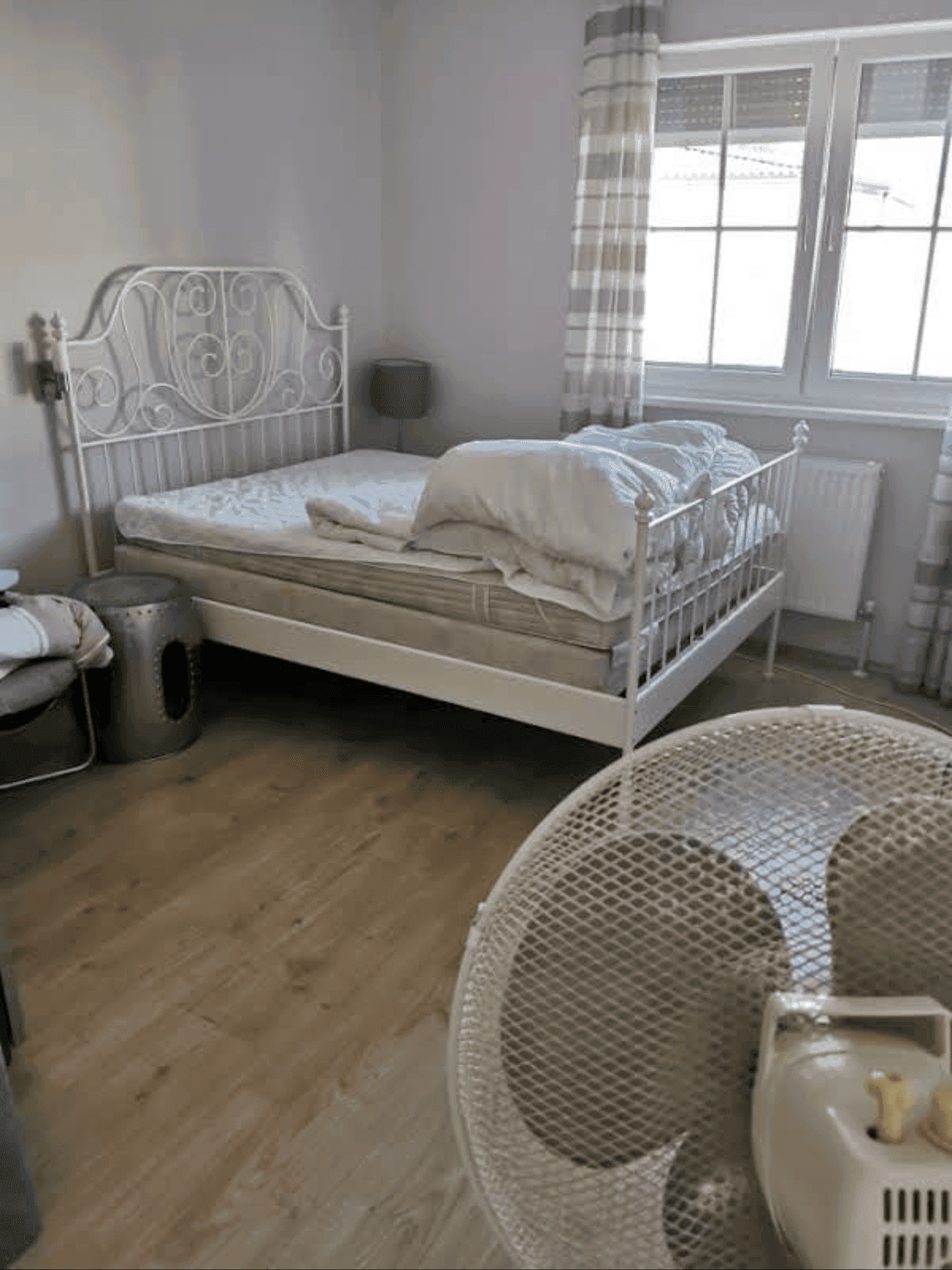 Schlafzimmer mit einem weißen Metallbett und einem Ventilator am Boden.