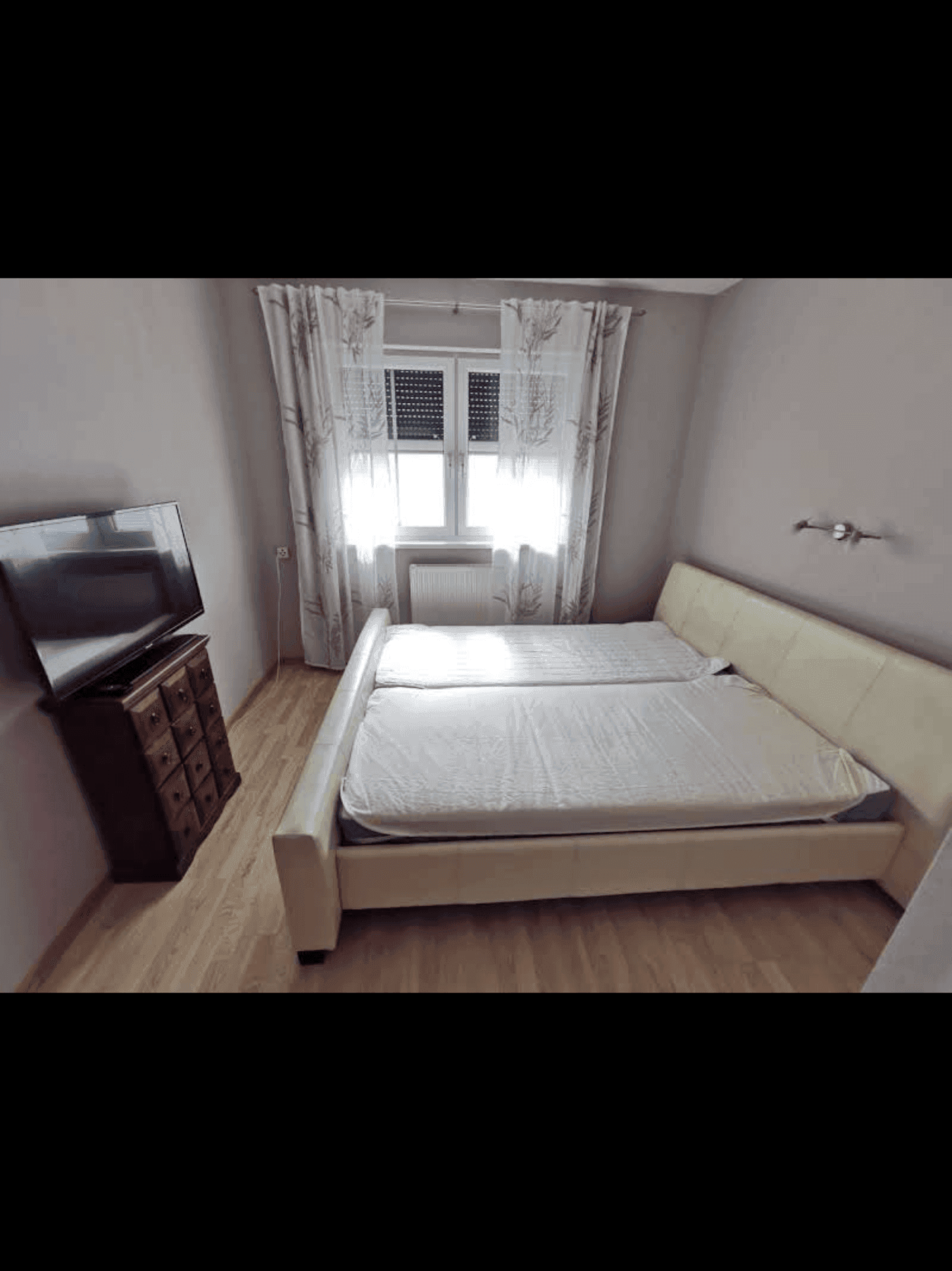 Gemütliches Schlafzimmer mit Doppelbett, Fernseher auf einer Kommode und Fenster mit Vorhängen.