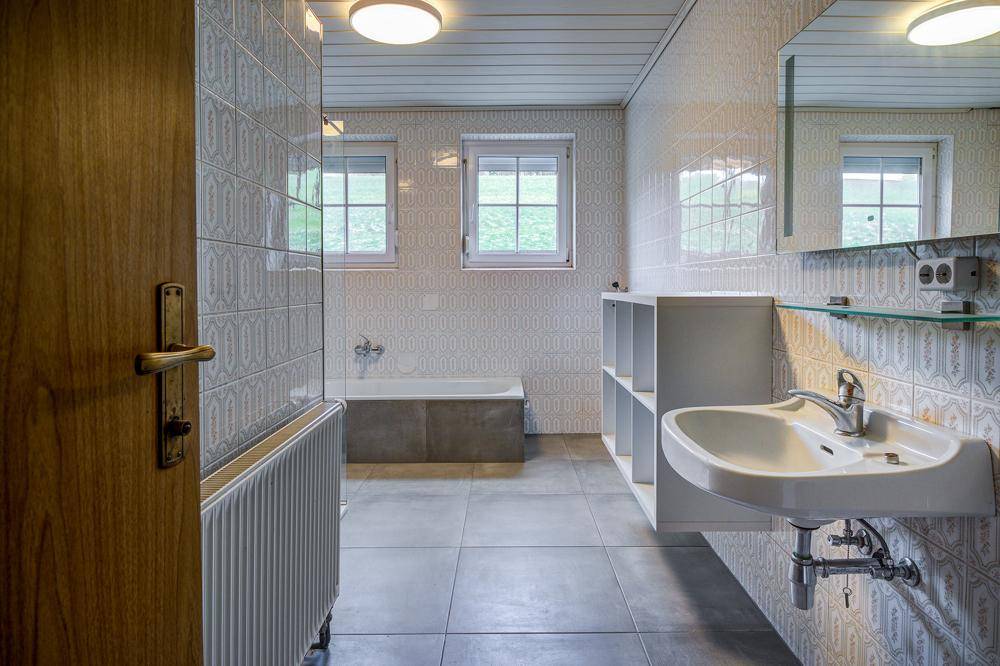 Badezimmer mit Badewanne, Waschbecken und gemusterten Fliesen, hell durch zwei Fenster.