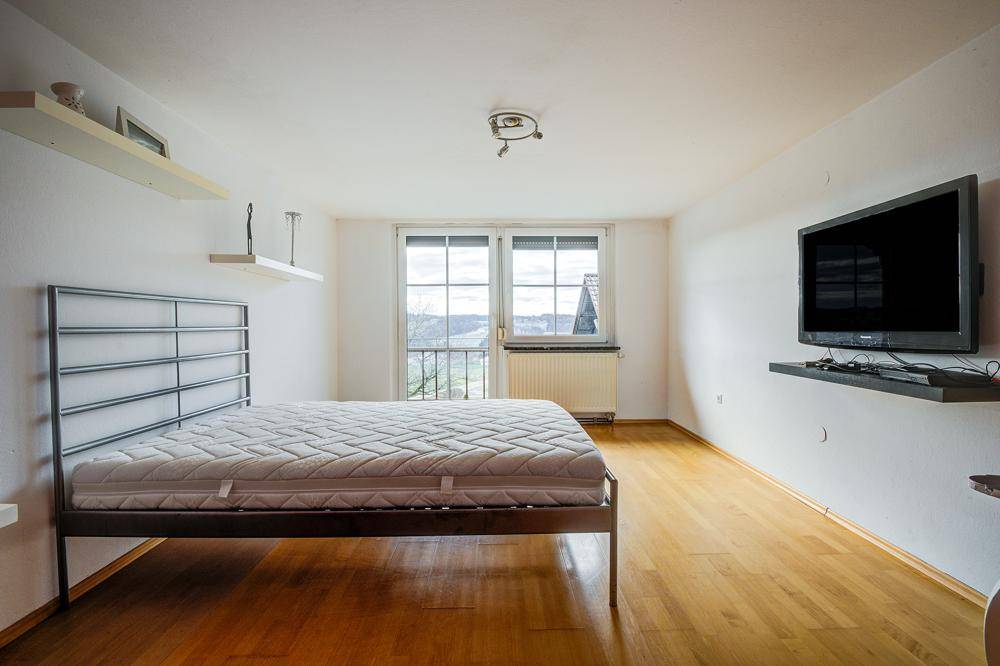 Helles Schlafzimmer mit Holzboden, Metallbett und Wandfernseher, mit Blick ins Grüne.