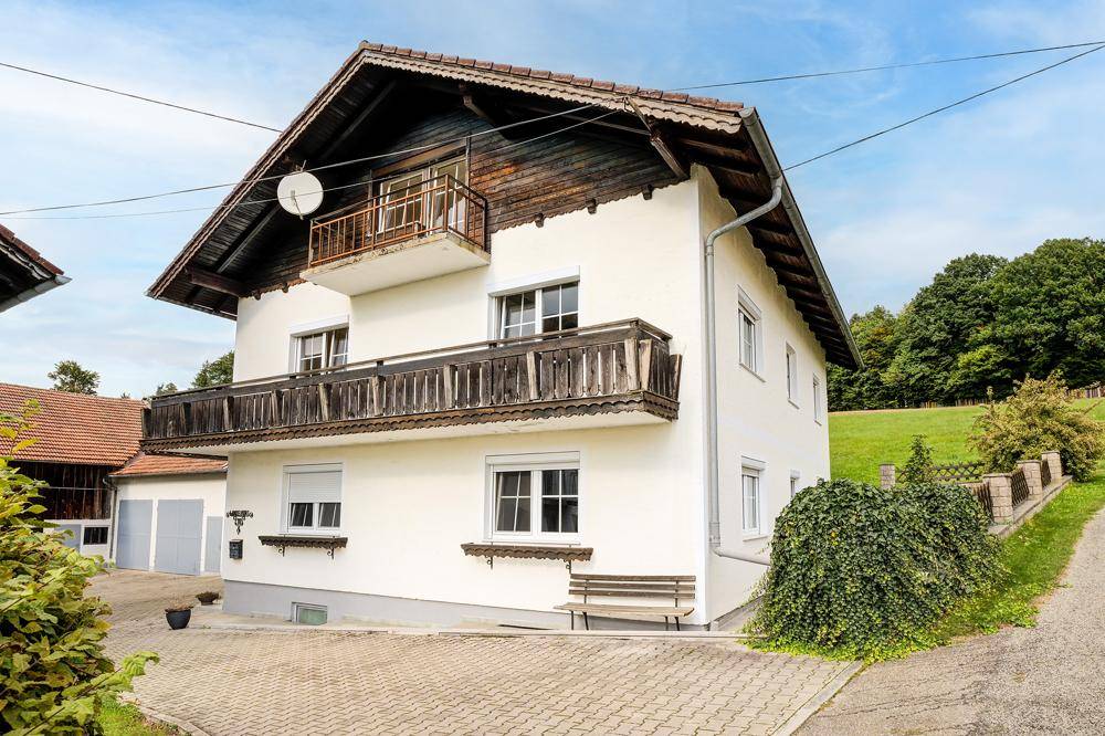 Weißes Wohnhaus mit traditionellen Holzbalkonen und gepflasterter Auffahrt, umgeben von Natur.