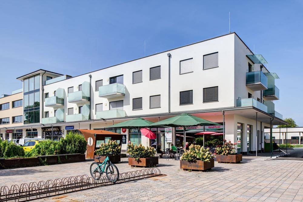 Modernes Gebäude mit Geschäften, Außengastronomie und Balkonen unter blauem Himmel.