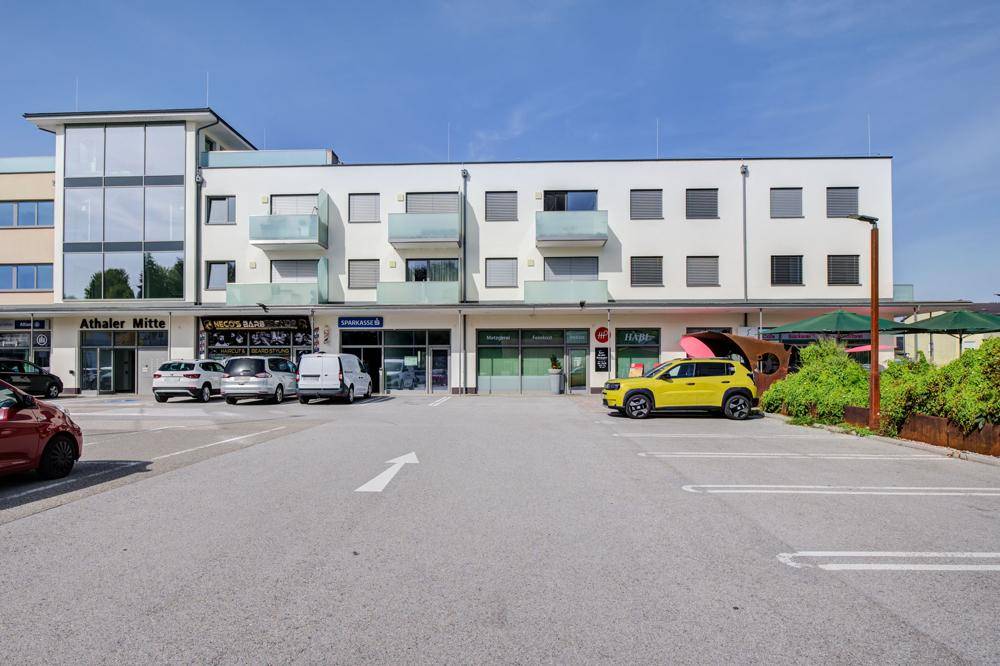 Modernes Geschäfts- und Wohngebäude mit Parkplätzen und Außengastronomie.
