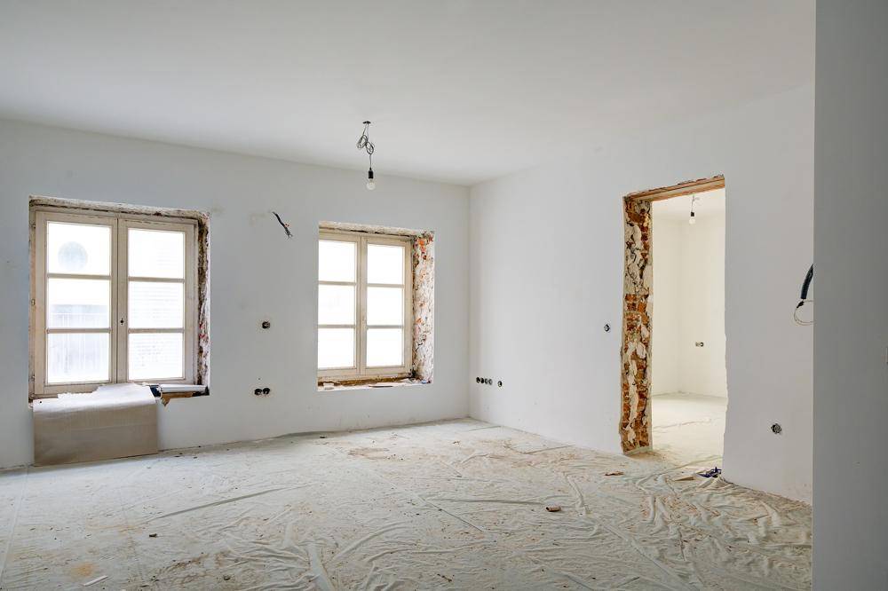 Unfertiger Innenraum mit zwei Fenstern und freigelegten Türrahmen, bereit für den Ausbau.