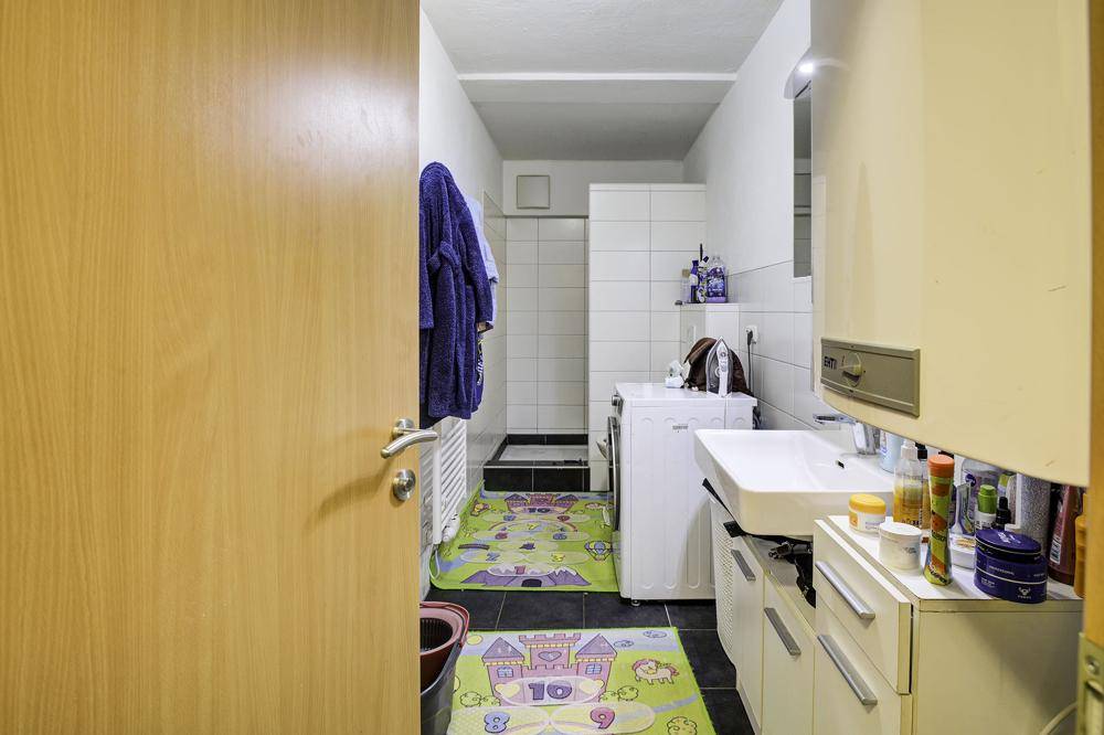 Badezimmer mit Dusche, Waschbecken und Waschmaschine, ausgestattet mit Fliesenboden und weißen Wänden.