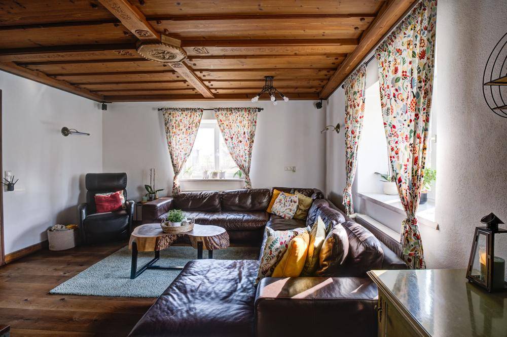 Gemütliches Wohnzimmer mit Holzbalkendecke, Ledersofa und bunten Vorhängen.