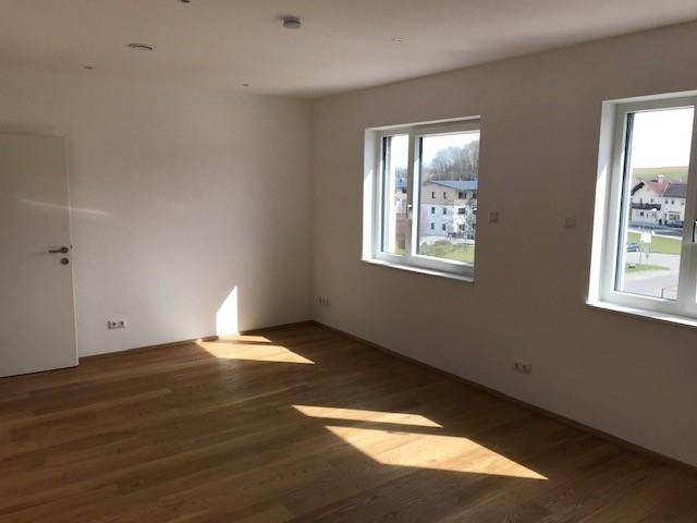 Lichtdurchfluteter Wohnraum mit Holzboden und zwei großen Fenstern, die viel Sonne hereinlassen.