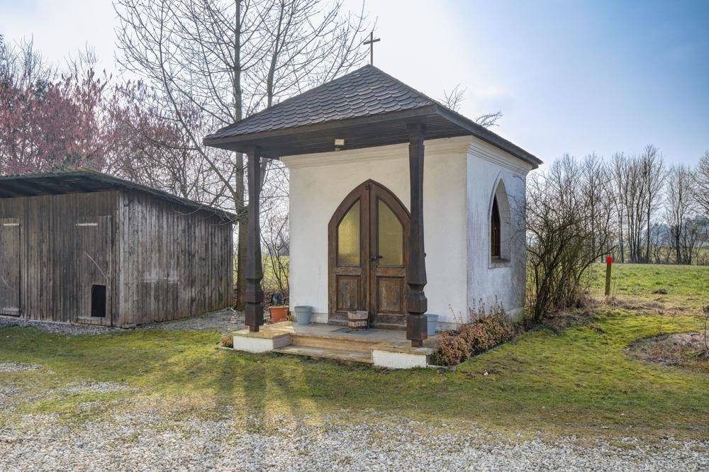 Kleine Kapelle mit Holztüren und Vordach, umgeben von Gras und Bäumen.
