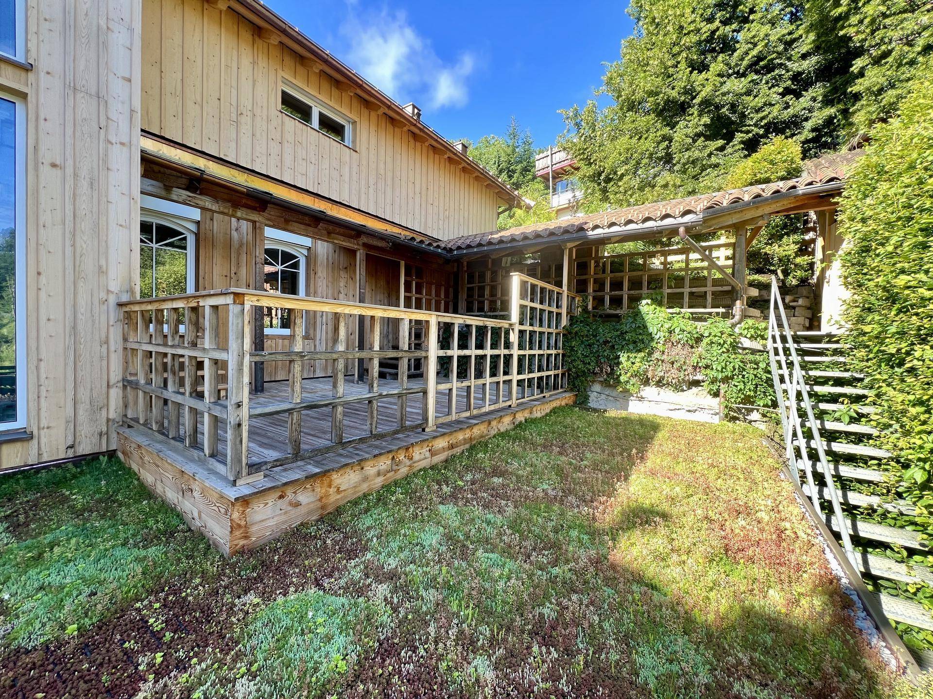 Holzterrasse mit Geländer und Treppe, umgeben von üppigem Grün und einer Holzfassade.