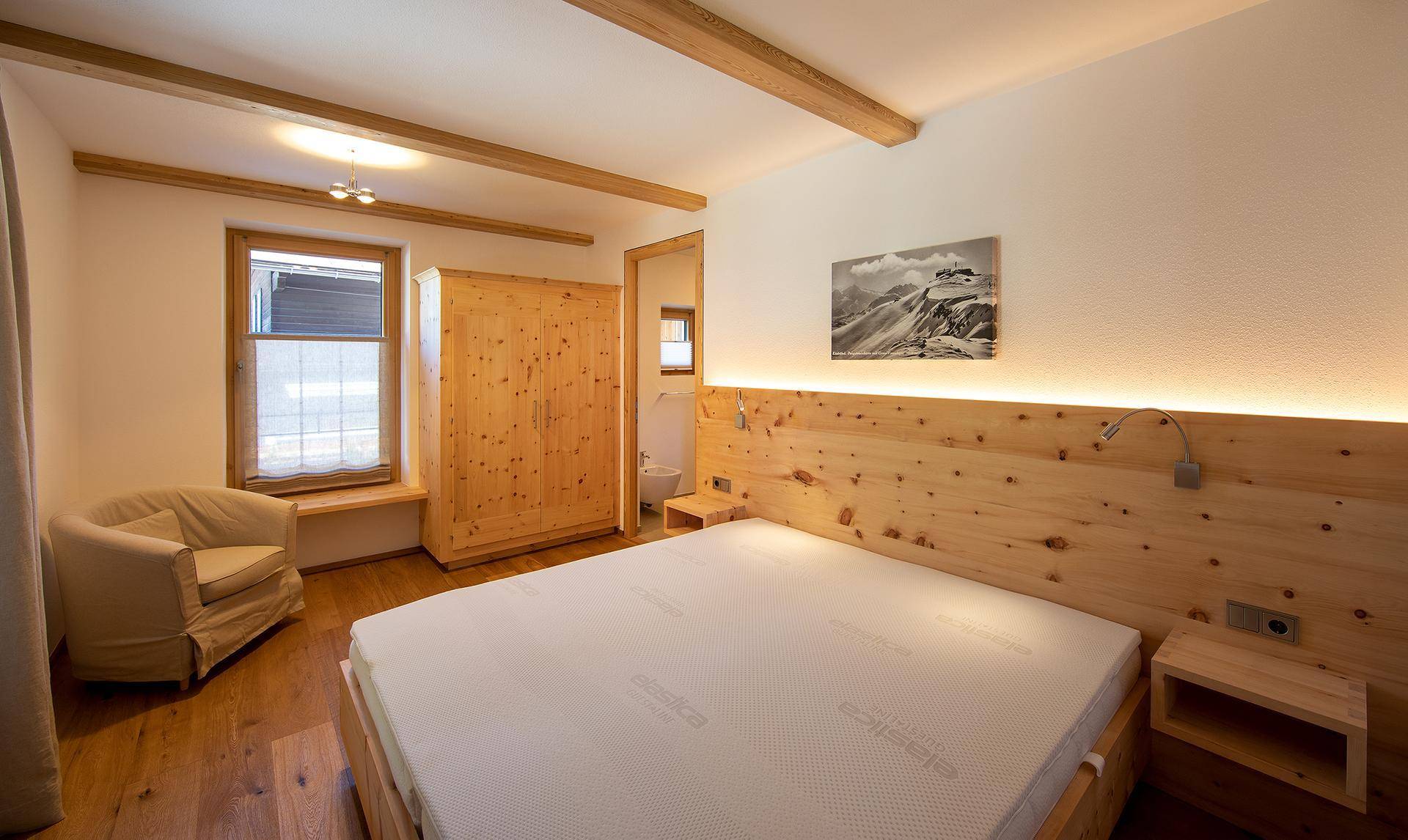 Helles Schlafzimmer mit Holzbett, Einbauschrank und Holzbalken, mit Fenster und Sitzgelegenheit.