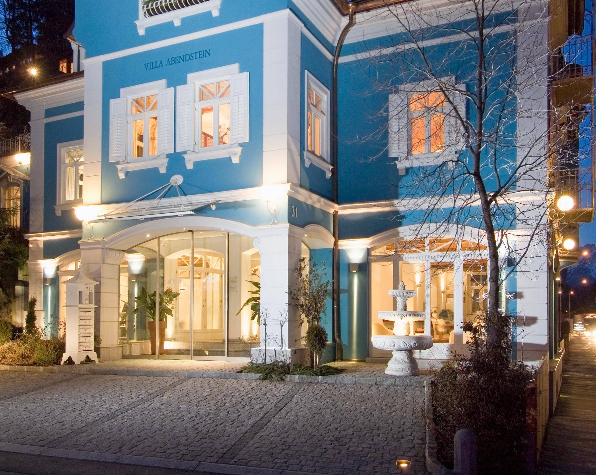 Beleuchtete blaue Villa bei Nacht mit dekorativem Brunnen und einladendem Eingangsbereich.