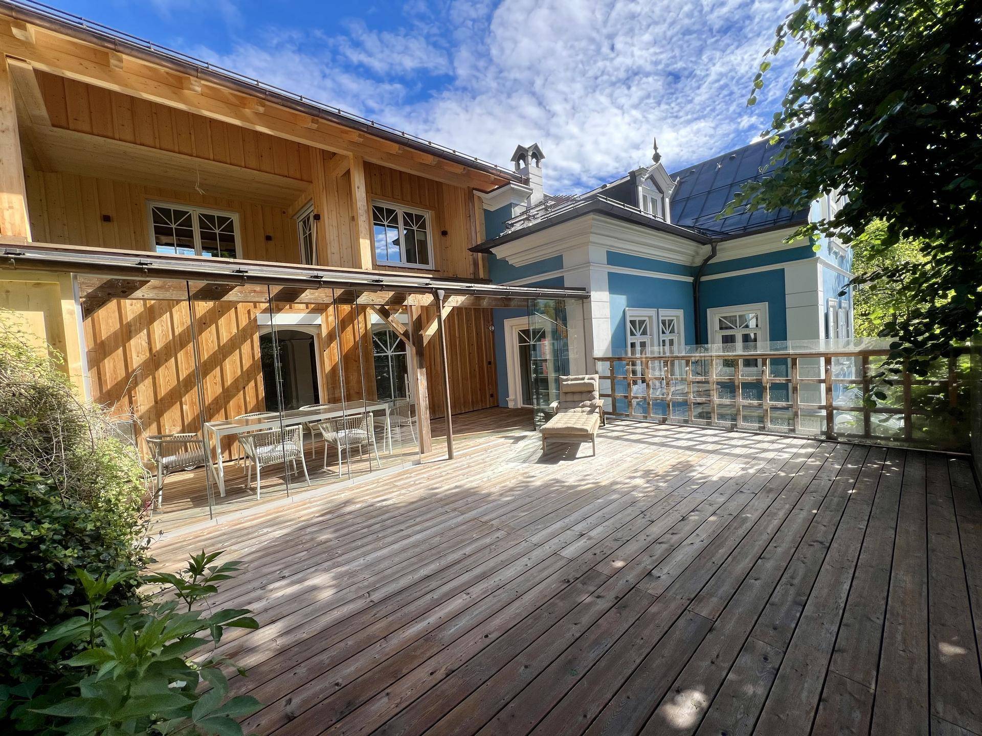 Geräumige Holzterrasse mit Sitzgelegenheiten und Blick auf die blaue Villa und die umliegende Natur.