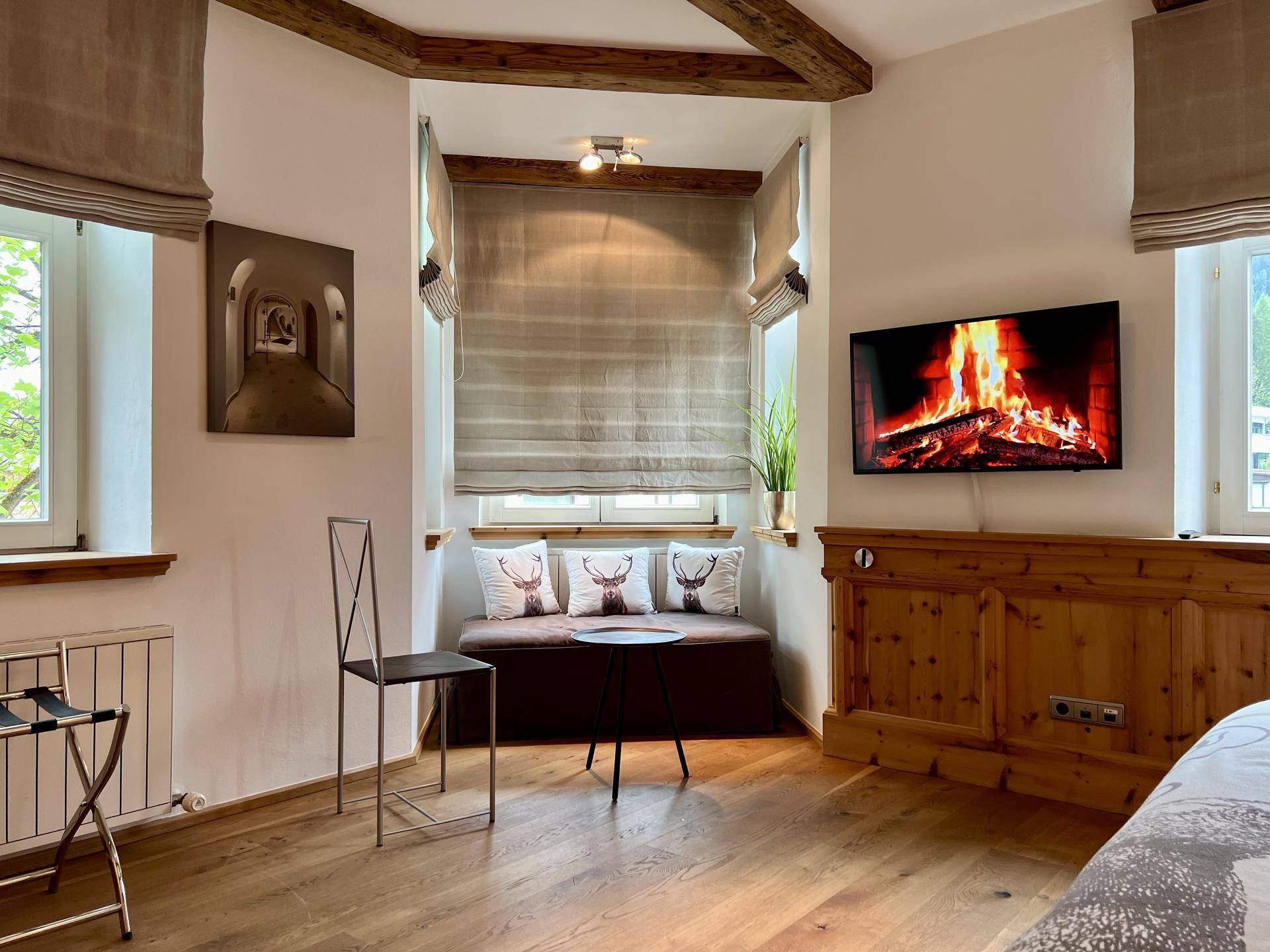 Schlafzimmer mit gemütlicher Fensternische, Holzboden und einem Fernseher, der ein Kaminfeuer zeigt.