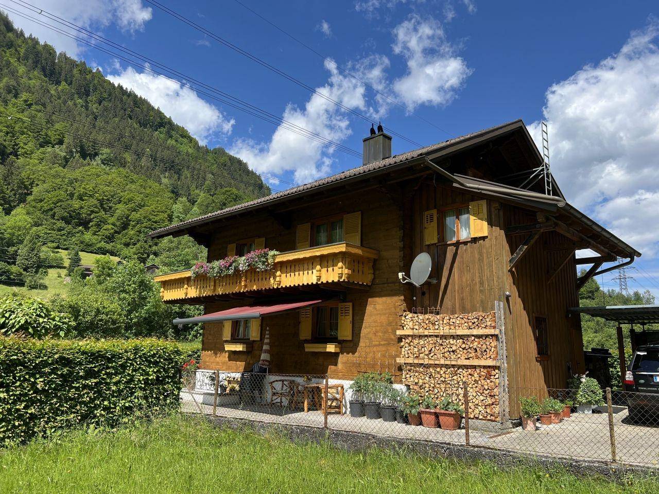 Seitenansicht des Holzhauses mit gestapeltem Brennholz und Blick auf die bewaldeten Berge.