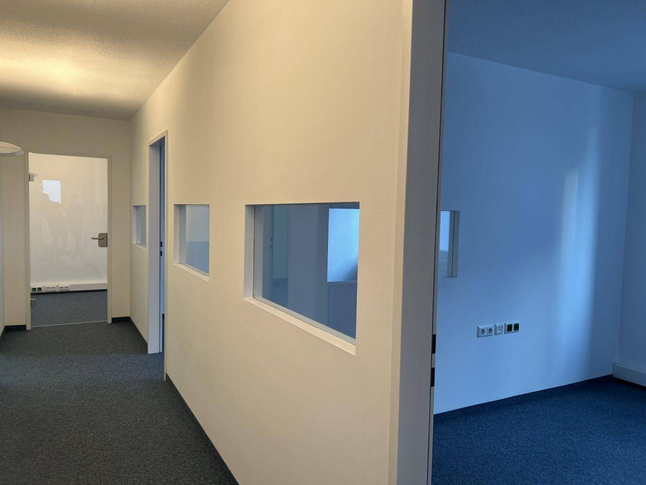 Heller Flur mit Teppichboden und kleinen Fenstern in der Trennwand zu angrenzenden Räumen.