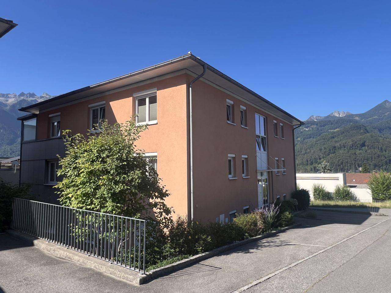 Seitenansicht des Gebäudes mit gepflegter Fassade und Parkmöglichkeiten, umgeben von Bergen.