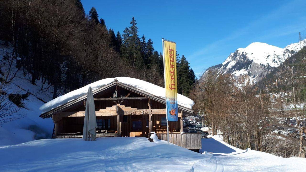 Traditionelles Holzgebäude im Schnee vor einer beeindruckenden Bergkulisse unter strahlend blauem Himmel.