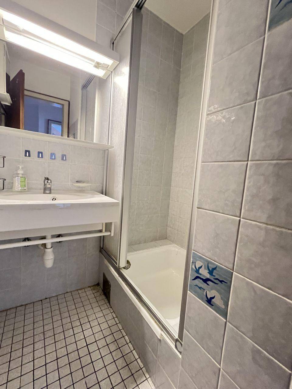 Badezimmer mit hellblauen Fliesen, Einzelwaschbecken und Badewanne mit Duschabtrennung.