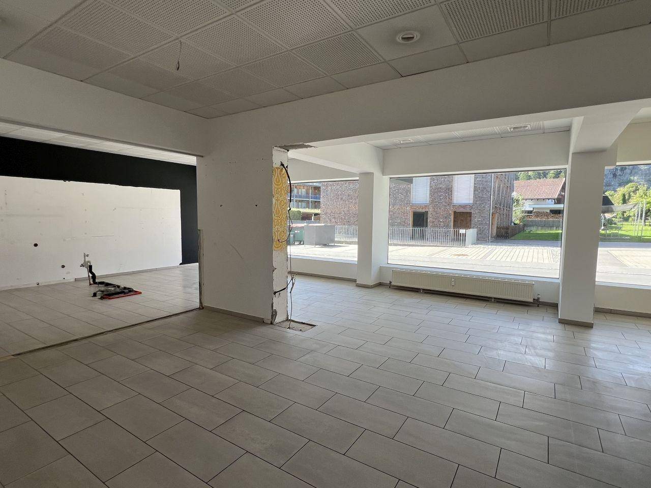 Heller, leerstehender Ladenraum mit Fliesenboden, abgehängter Decke und großen Fenstern zur Straße hin.