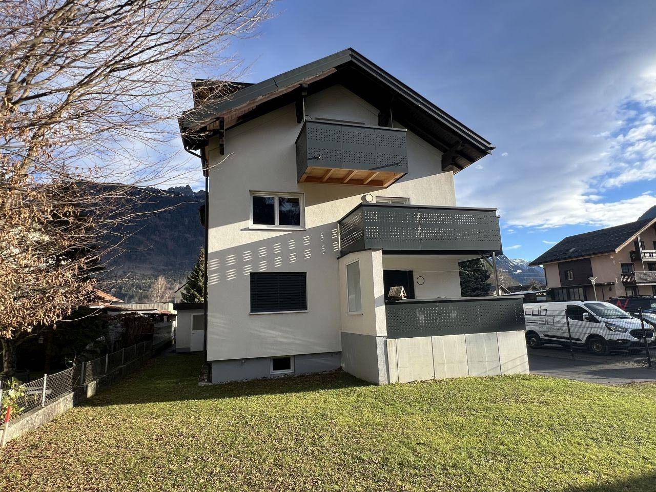 Außenansicht des modernen Gebäudes mit Balkonen, Gartenfläche und Bergblick im Hintergrund.